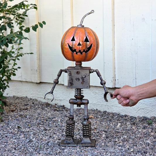 A hand is holding a pumpkin-shaped metal sculpture with a carved face, standing on a gravel surface next to a white wall.