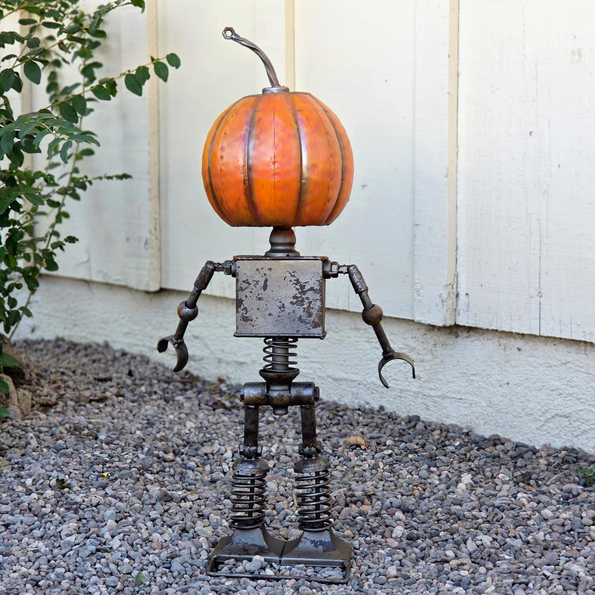 A metal pumpkin-shaped sculpture stands on a gravel surface, with a white wooden wall in the background.