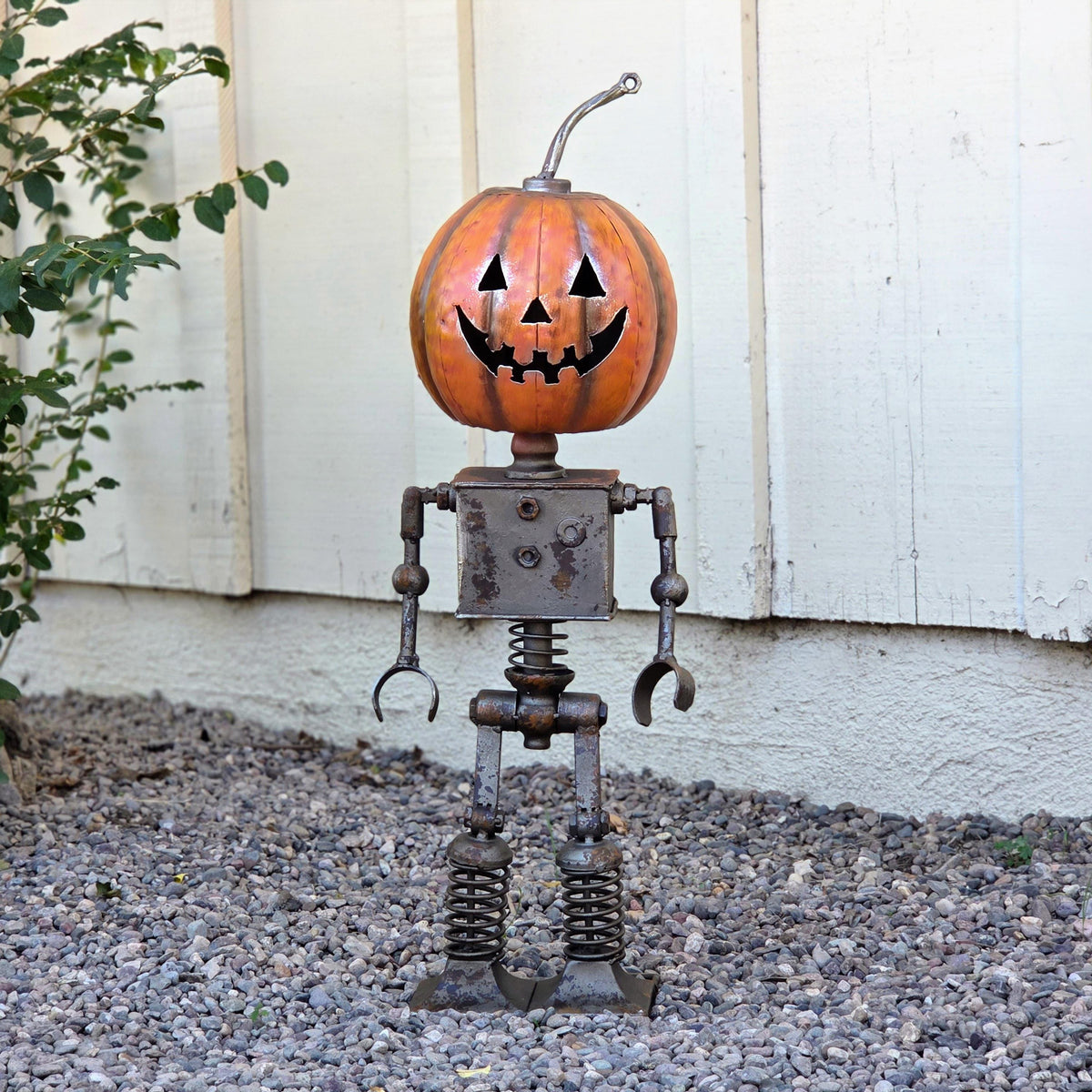 A Halloween-themed metal pumpkin robot sculpture stands on a gravel surface, with a pumpkin head and a carved face.
