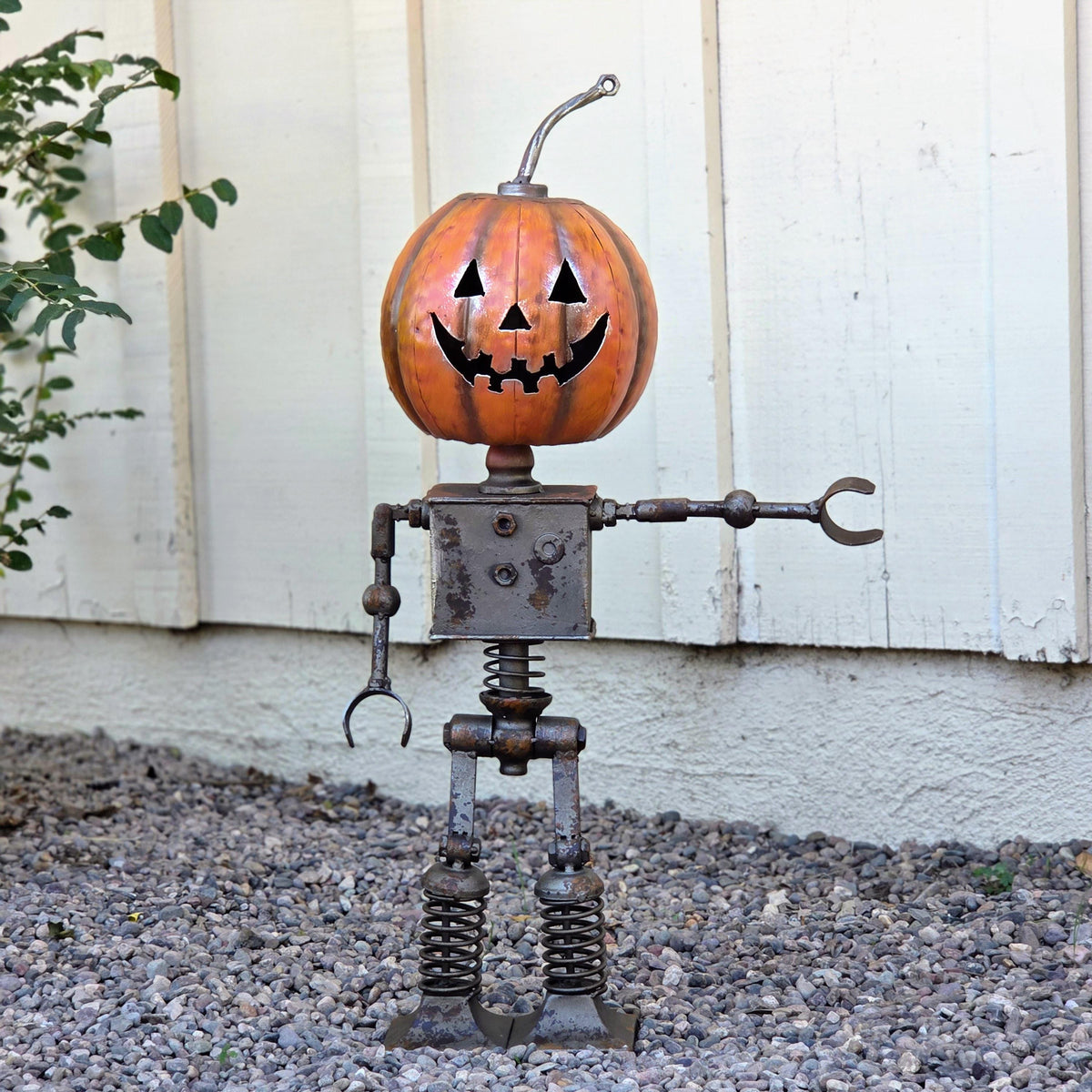 A metal pumpkin-shaped robot sculpture stands on a gravel surface, with a white wooden wall in the background.