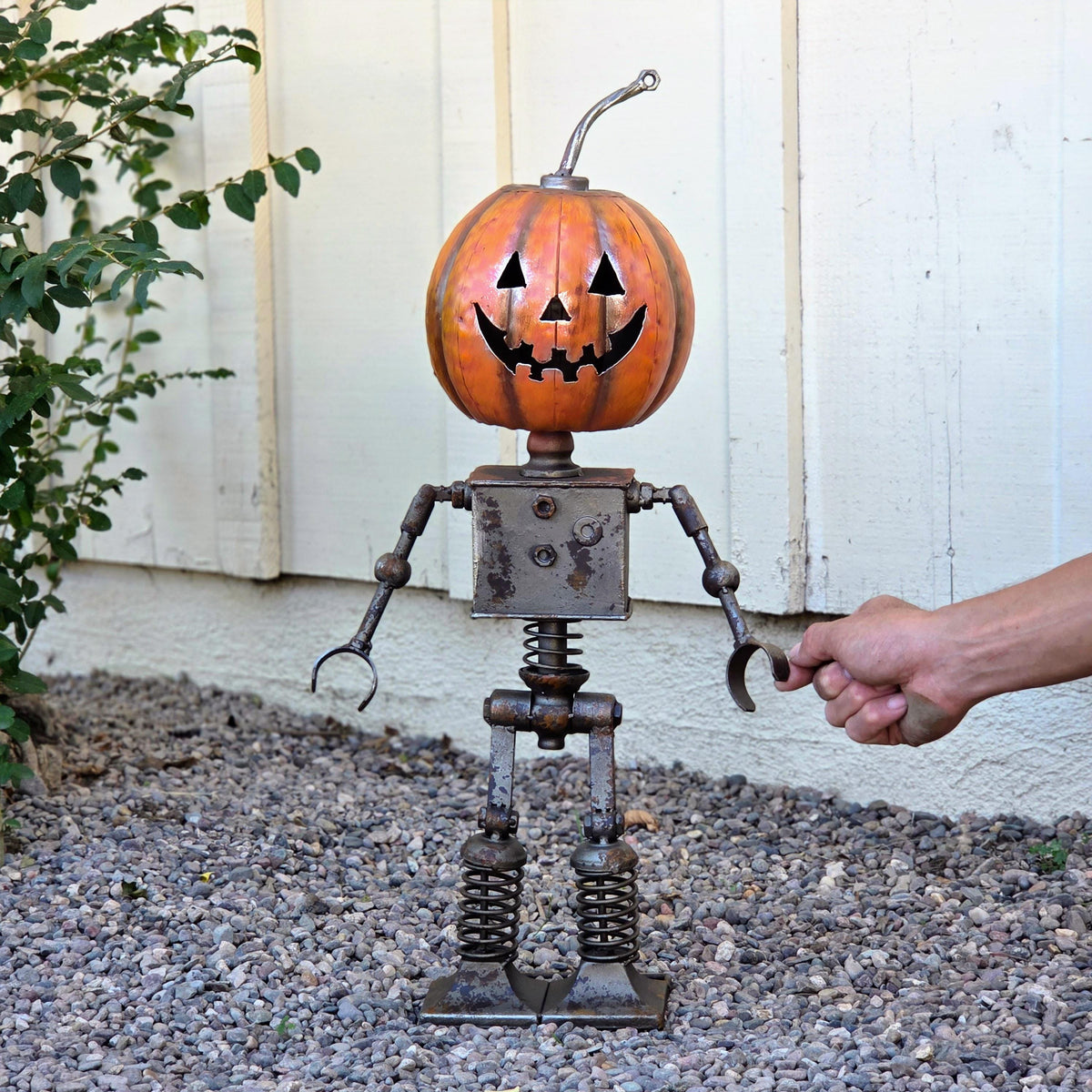 A hand is holding a pumpkin-shaped metal sculpture with a carved face, standing on a gravel surface next to a white wall.