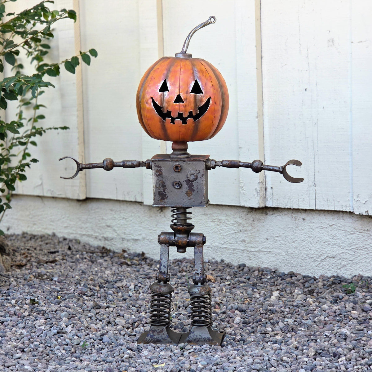 A Halloween-themed metal pumpkin robot sculpture stands on a gravel surface, with a pumpkin head and a carved face.